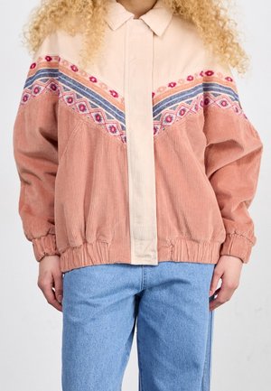 Veste en velours côtelé avec poignets et taille élastiques, présentant un motif géométrique multicolore sur la poitrine, portée avec un jean bleu clair.