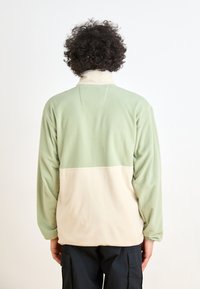 Pullover di pile verde chiaro e crema con colletto alto, caratterizzato da una superficie texturizzata e maniche lunghe, indossato con pantaloni cargo neri.