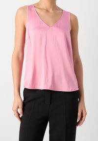 Woman wearing sleeveless pink V-neck blouse and black pants, standing against a plain light background.