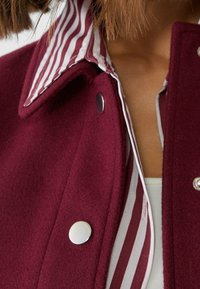Burgunderfarvet uldjakke med en sølvfarvet trykknaplukning og en rød-hvid stribet krave, der har en blød tekstur og skræddersyet design.