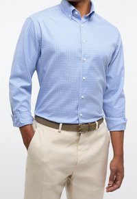 Modrá košile s puntíkovaným vzorem gingham, límcem na knoflíky, dlouhými rukávy vyhrnutými na manžetách, v kombinaci s béžovými zúženými kalhotami a texturovaným hnědým páskem.