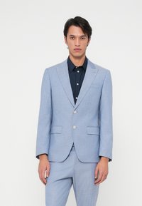 Šviesiai mėlynas švarko švarkas su tekstūruotu paviršiumi, vieno mygtuko dizainas, išpjovos apykaklė ir dvi priekinės kišenės, dėvimas ant tamsios marškinėlių.