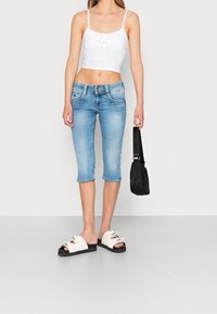 Jeans cortos azul claro con un diseño ajustado, combinados con un top corto blanco y sandalias blancas con suelas negras y lazos. Bolso negro en la mano.