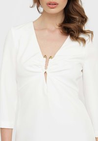Abito bianco con scollatura a V, dettaglio arricciato sul petto e accenti dorati. Maniche lunghe e texture del tessuto liscia, design aderente.