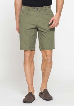Uomo che indossa pantaloncini verde oliva fino al ginocchio e scarpe slip-on marrone scuro in piedi davanti a uno sfondo neutro.