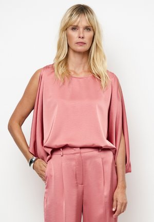 Blonde vrouw die een losse, mouwloze roze satijnen blouse draagt met bijpassende hoge taille broek, staand met één hand in haar zak.