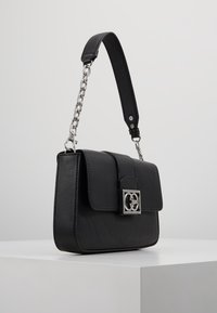 Black leather handbag with silver chain and leather strap, rectangular shape, front flap, and silver decorative clasp on white pedestal.