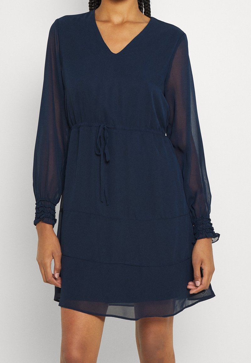 Femme portant une robe bleu marine à manches longues, col en V, taille à cordon et poignets élastiques, debout devant un fond uni.