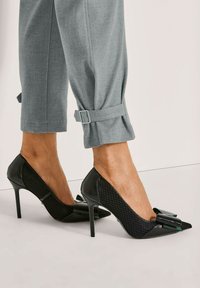 Benen in grijze tapered broek met enkelriemen en zwarte hoge hakken met gestructureerde zijkanten en decoratieve strikken op de spitse neuzen.