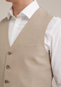 Beige Weste aus strukturiertem Stoff, mit V-Ausschnitt, fünf braunen Knöpfen und einer Brusttasche, getragen über einem weißen Hemd.