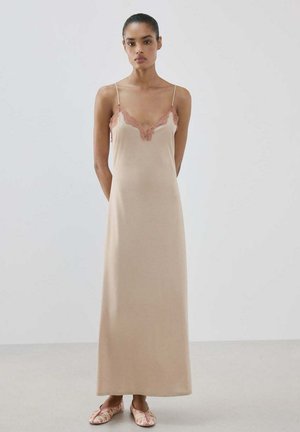 Femme portant une robe nuisette beige avec un bord en dentelle et des ballerines assorties, debout devant un fond gris clair uni.