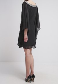 Robe en chiffon noir au design fluide, avec un col orné de accents argentés et des manches longues. Associée à des talons hauts noirs.