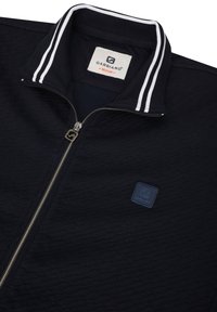 Jūrus laivyno zip-up striukė su tekstūruotu audiniu, balti dryžiai ant apykaklės, logotipo emblema ir sidabro spalvos užtrauktuko detalės.