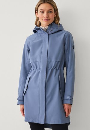 Vrouw met een lichtblauwe regenjas met capuchon, voorzakken en een verstelbare taille, staand tegen een effen lichtgrijze achtergrond.