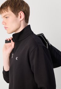 Čierna zateplená bunda na zips s vysokým golierom, s malým bielym logom na hrudi, doplnená čiernou taškou, ktorú drží na jednom ramene.