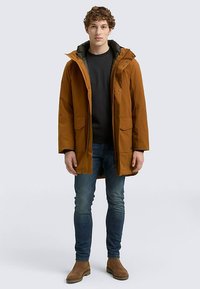 Hnědá parka s kapucí, přední zapínání na patentky, dvě velké kapsy, nošena přes černé tričko a modré džínsy. Outfit doplňují hnědé boty.