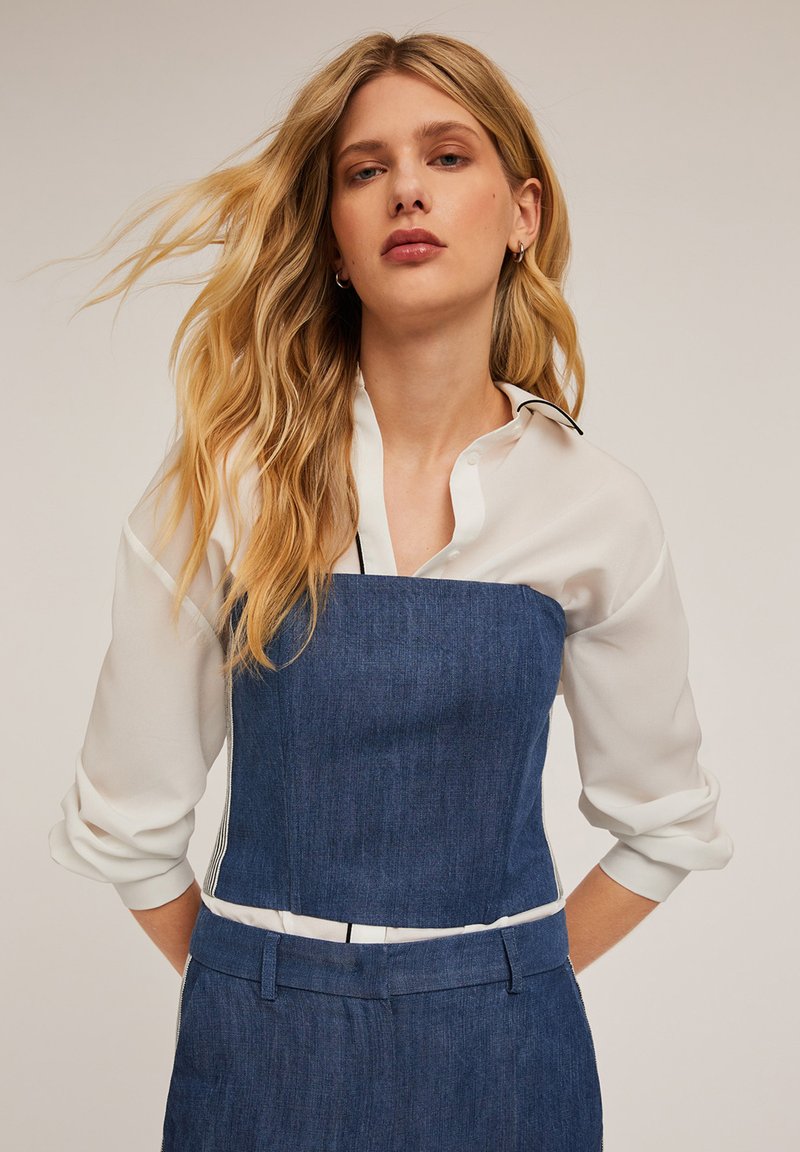 Jeansklänning med en vit blus under. Har en strukturerad livstycke, långa ärmar och kontrasterande svart piping längs kragen.