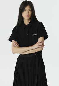 Zwarte poloshirt met een knoopsluiting bij de kraag en korte mouwen, voorzien van een wit logo op de borst. Gestyled met een geplooide zwarte rok.
