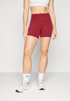 Kobieta w burgundowych sportowych szortach z wysokim stanem, białym krótkim topie, białych skarpetach Nike oraz białych butach sportowych, stojąca na jednolitym tle.