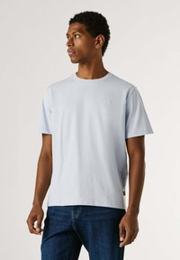 Homme portant un T-shirt à manches courtes bleu clair et un jean bleu foncé, regardant vers sa gauche sur un fond clair uni.