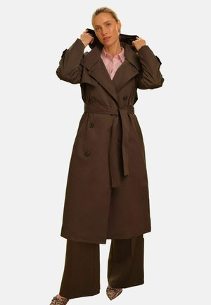 Woman adjusting the hood of a long dark brown belted trench coat over wide-leg pants and leopard print shoes, against a plain background.