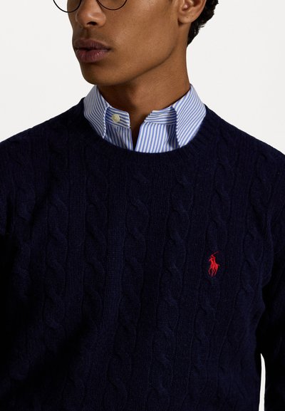 Marineblauer Kabelstrickpullover aus strukturiertem Stoff, mit Rundhalsausschnitt und einem kleinen roten gestickten Logo auf der Brust, über einem gestreiften Hemd getragen.
