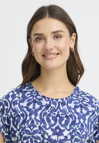 Sinine ja valge trükimustriga pluus keeruka lillemustriga. Pehme kangas, ümmargune kaelus, lühikesed varrukad, esile tõstetud väikeste rõngaskõrvarõngastega.