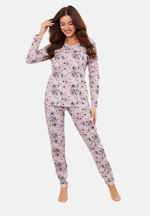 Kobieta uśmiechnięta, ubrana w jasnoróżową, kwiatową bluzkę z długim rękawem i pasujące spodnie, stoi boso na jednolitym tle.