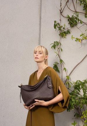 Blonde woman in olive dress holding large dark brown woven leather handbag stands against light wall with green climbing plants.
