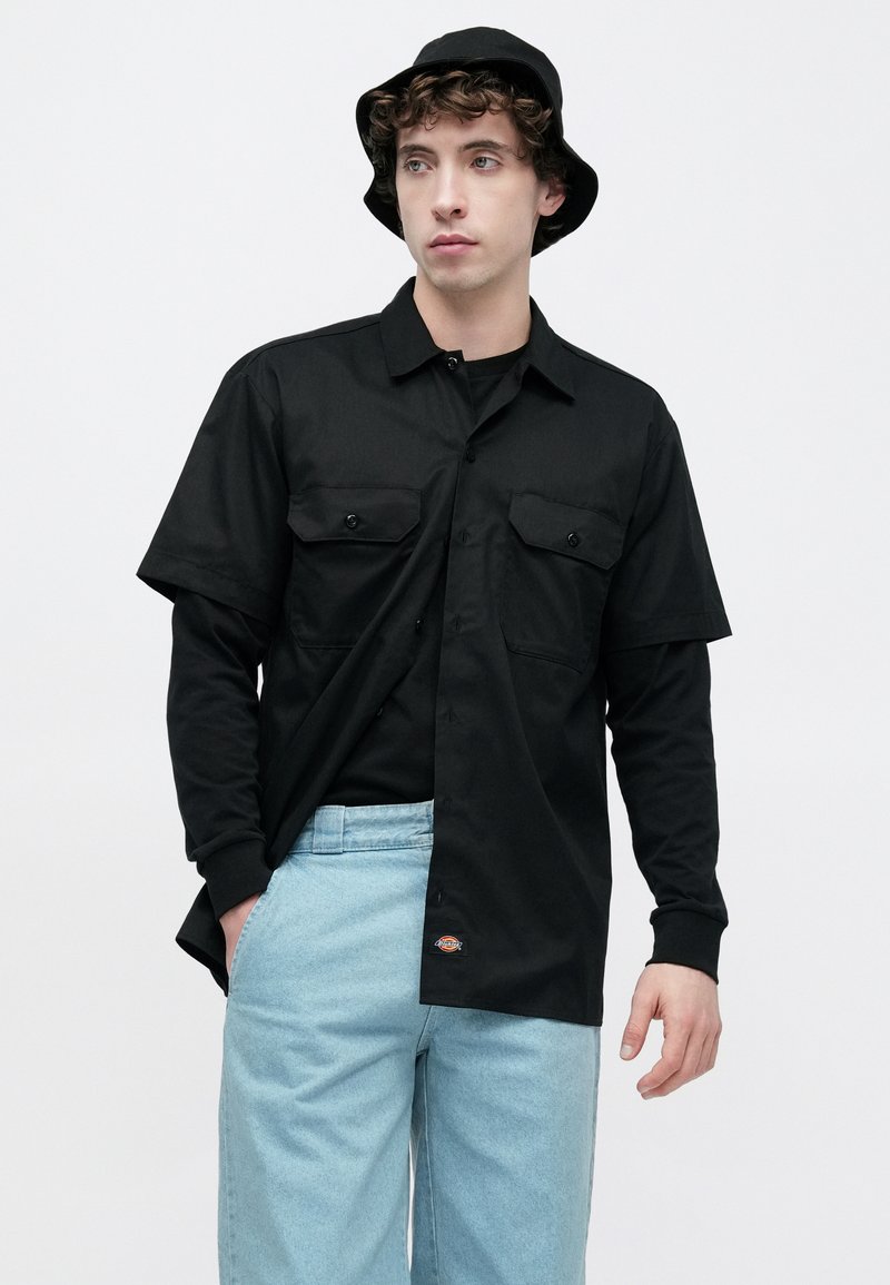 Chemise noire à manches courtes avec deux poches avant, portée sur une chemise noire à manches longues. Pantalon en denim bleu clair et chapeau seau noir.