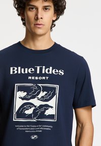 Tamsiai mėlynos spalvos medvilninis marškinėliai su balta iliustracija, vaizduojančia bangas, ir užrašu „Blue Tides Resort“ priekyje. Klasikinė apvalios kaklo išpjova.