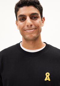 Zwarte sweatshirt met een ronde hals, voorzien van een klein geel geborduurd logo aan de linkerkant. Soepele stoftextuur.