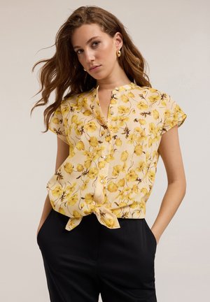 Short-sleeve, button-up blouse in soft yellow fabric adorned with floral patterns, featuring a tied front and relaxed fit.