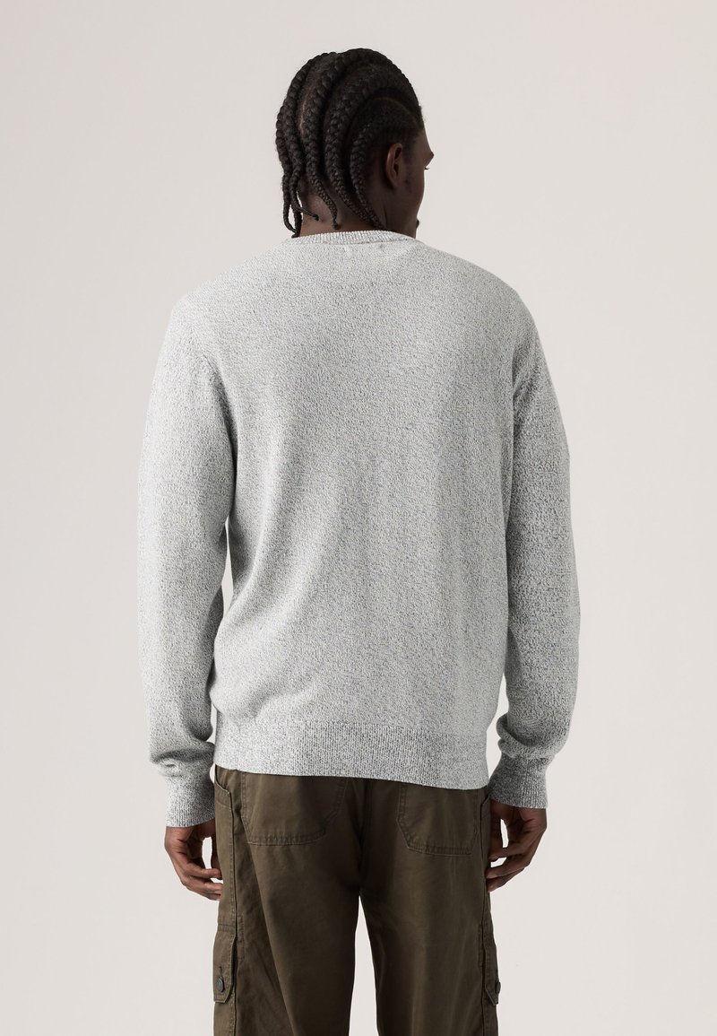 Mann mit geflochtenen Haaren, der einen hellgrauen, strukturierten Pullover und olivgrüne Cargohosen trägt, steht mit dem Rücken zu einem schlichten Hintergrund.