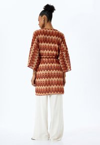 Lång, mönstrad kimono i röd, orange och krämfärgad zigzagdesign, med trekvartsärmar och knytmidja, tillsammans med lösa vita byxor.