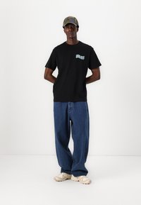 Zwart katoenen T-shirt met korte mouwen en een blauw grafisch logo op de borst, gecombineerd met losse blauwe spijkerbroeken en beige sneakers.