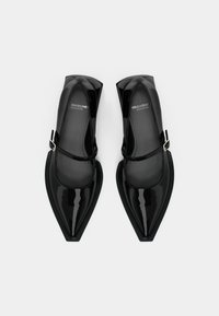 Paire de chaussures Mary Jane noires en cuir verni à bout pointu, avec brides à la cheville et talons carrés sur fond gris clair.