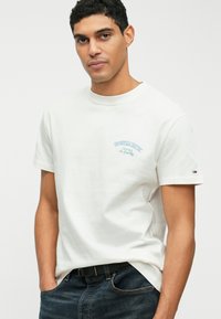 Tricou din bumbac alb cu logo texturat albastru pe pieptul stâng, guler rotund și mâneci scurte. Asortat cu blugi denim închis.