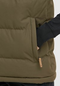 Gilet matelassé vert avec une surface texturée, présentant une poche latérale et une étiquette en tissu. À l'intérieur, on aperçoit une chemise à manches longues noire.