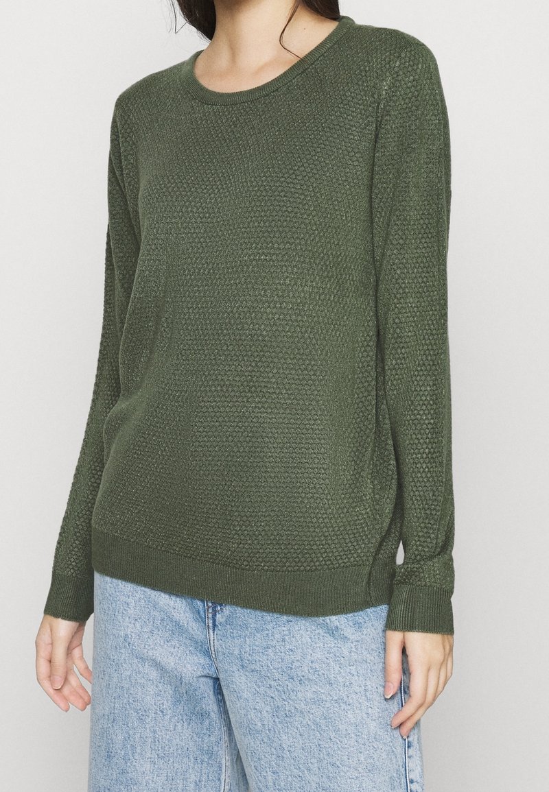 Grüner Strickpullover mit strukturiertem Muster, Rundhalsausschnitt und langen Ärmeln, kombiniert mit hellblauen Jeanshosen.