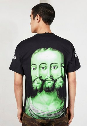 Mężczyzna zwrócony tyłem, ubrany w czarny t-shirt z dużym zielonym artystycznym nadrukiem twarzy brodatego mężczyzny pokazującej trzy nachodzące na siebie głowy.