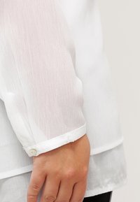 Camisa blanca, ligera y de manga larga con puños abotonados; cuenta con una tela texturizada y un sutil diseño en capas en el dobladillo.