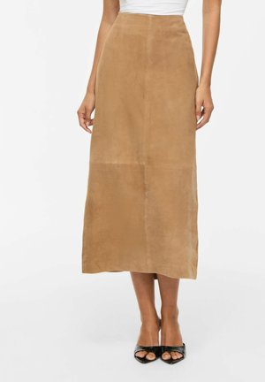 Femme portant une jupe midi taille haute en daim couleur fauve associée à des talons ouverts noirs et un haut blanc.