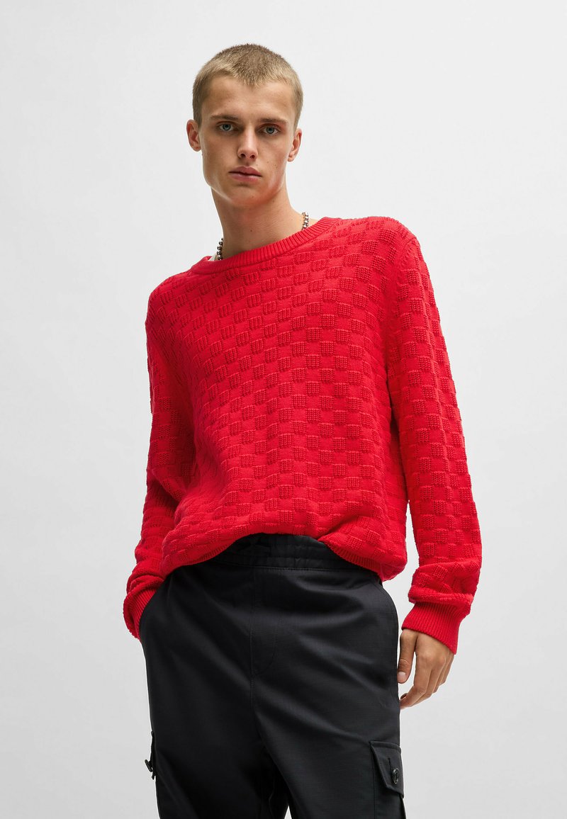 Maglione rosso lavorato a maglia con un motivo a quadri testurizzato e polsini a costine. Abbinato a pantaloni neri con tasche laterali.