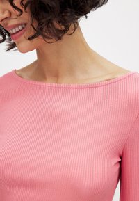 Rozā ribota blūze ar plašu kakla izgriezumu, izceļot mīkstu faktūru un pieguļošu dizainu. Audums šķiet nedaudz spīdīgs.