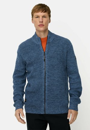 Man wearing a blue knitted zip-up sweater over an orange shirt standing against a plain background.