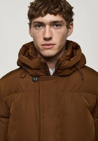 Brown puffer jacket featuring a high collar, zippered front, adjustable drawstring hood, and button accents. Smooth texture with quilted design.