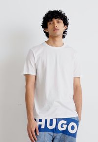Camiseta blanca de algodón con cuello redondo, mangas cortas, combinada con pantalones azules con estampado que presentan letras en negrita.