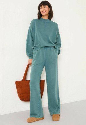 Übergroßer, türkisfarbener Baumwoll-Sweatshirt, kombiniert mit passender, weiter Hose. Eine braune, gesteppten Tragetasche und beiges Schuhwerk vervollständigen den Look.