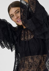 Melna mežģīņu topa ar volānu piedurknēm un caurspīdīgu rakstainu dizainu, ar vienkrāsainu melnu korseti zem tā, uzsverot smalku dizainu.